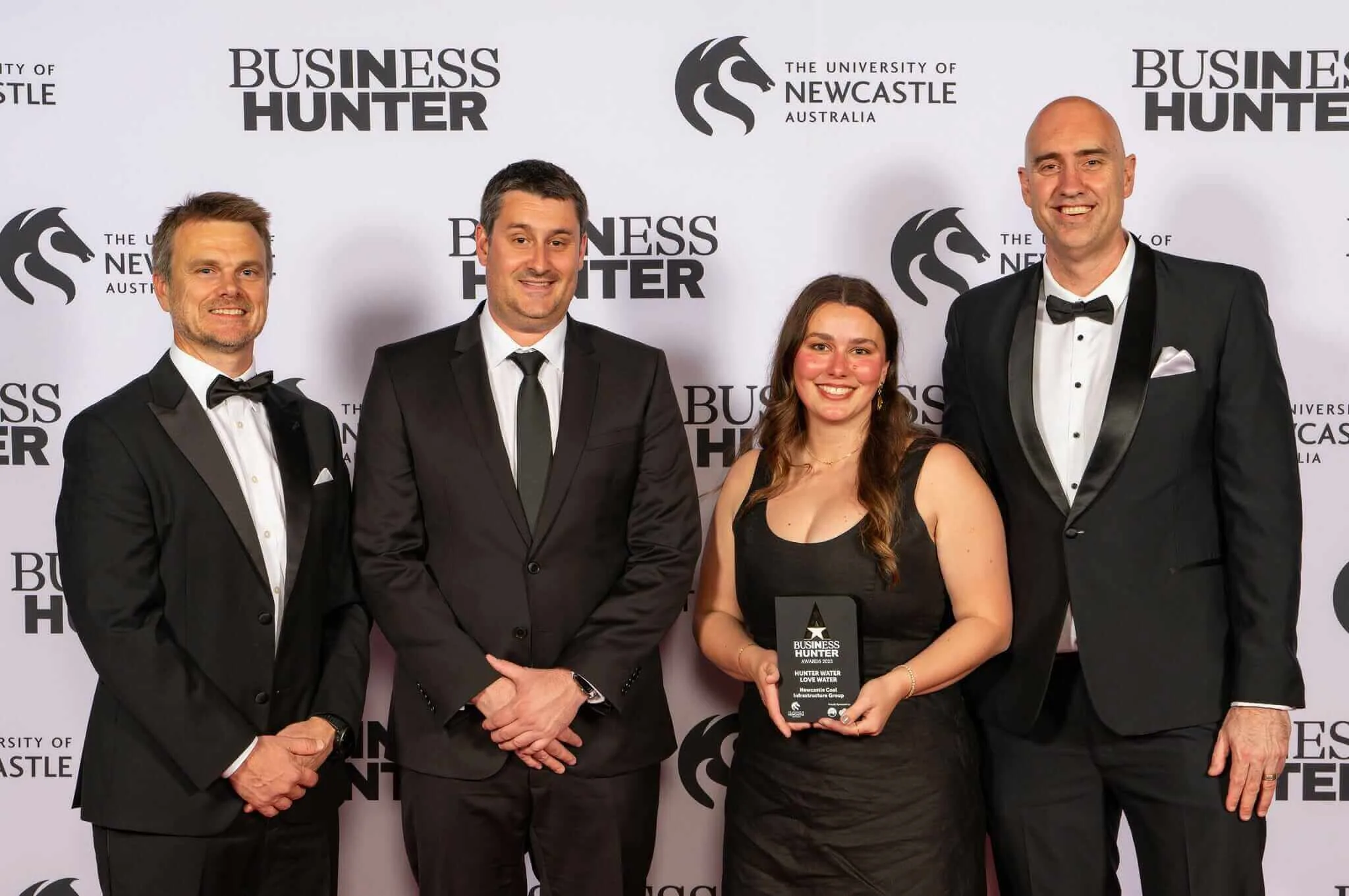 Three men in black suits and a lady in a black dress stand in front of a media wall with an award.